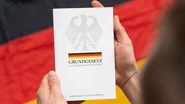 Mann hält Grundgesetz-Buch in der Hand vor deutscher Flagge | Bild: picture alliance / CHROMORANGE | Michael Bihlmayer