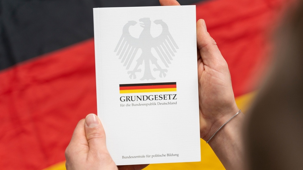 Mann hält Grundgesetz-Buch in der Hand vor deutscher Flagge | Bild: picture alliance / CHROMORANGE | Michael Bihlmayer