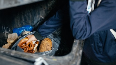 Eine Frau fischt Lebensmittel aus einer Mülltonne eines Supermarkts. | Bild: BR / Julia Müller