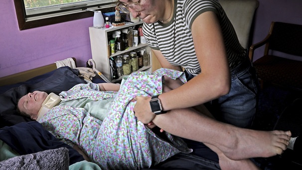 Deborah Smith wird von ihrer Helferin vom Rollstuhl aus auf das Bett gehoben. Sie leidet an ME/CFS. Deborah Smith hat ihre Wohnung in den USA schon über zwei Jahre lang nicht mehr verlassen können. | Bild: picture-alliance/dpa