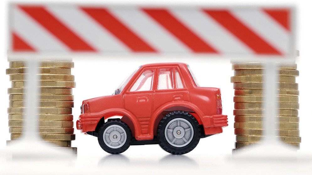 Ein Spielzeugauto auf zwei Stapeln Münzen unter einem Baustellen-Schild. | Bild: picture-alliance/dpa