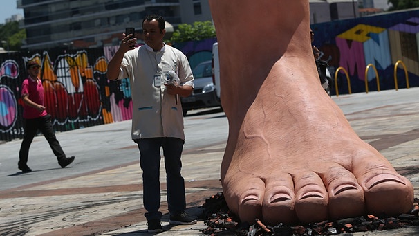 Sao Paulo: Ein Mann steht neben der riesigen Skulptur eines Fußes, der auf heißen Kohlen steht. Die Skulptur des brasilianischen Künstlers Eduardo Srur soll auf die Erbkrankheit Polyneuropathie aufmerksam machen, die sich unter anderem darin äußern kann, dass man Temperatur nicht mehr richtig fühlen kann. | Bild: picture-alliance/dpa