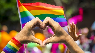 Hände formen vor einer Fahne mit Regenbogenfarben ein Herz. | Bild: stock.adobe.com/lazyllama