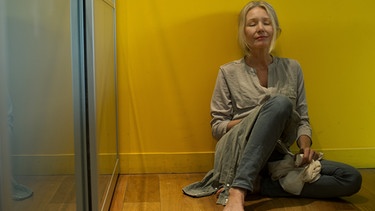 Eine barfüßige Frau sitzt erschöpft und depressiv vor einer gelben Wand und hat die Augen geschlossen. | Bild: picture-alliance/dpa