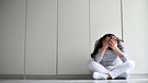 Eine junge Frau sitzt verzweifelt vor einer weißen Wand und stützt ihren Kopf mit den Armen. | Bild: picture-alliance/dpa