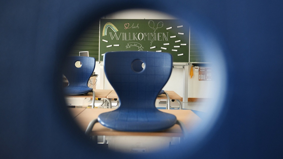 Symbolbild: Blick durch das Loch in der blauen Lehne eines Schulstuhls auf die nächste Stuhlreihe. Im Hintergrund eine Schultafel, auf der mit Kreide geschrieben "Willkommen" steht. | Bild: dpa-Bildfunk/Angelika Warmuth