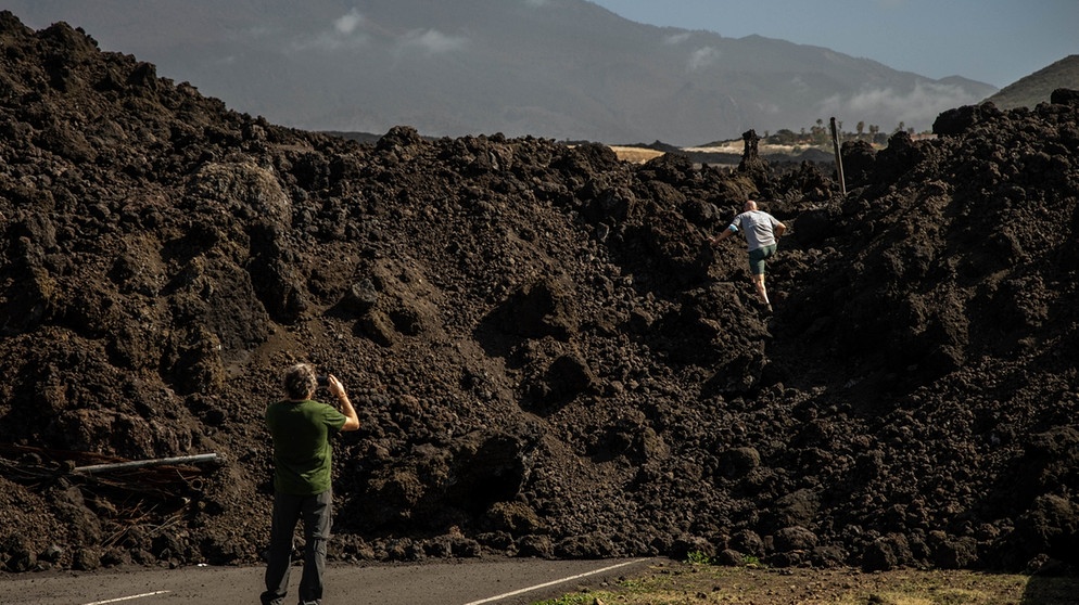 Ein Mann klettert auf einen meterhohen, versteinerten Lavastrom auf einer Straße in Tazacorte, während ein anderer ein Foto macht | Bild: dpa-Bildfunk/Kike Rincón