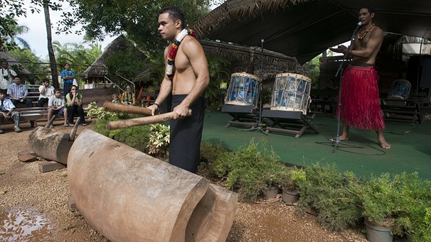 Ein Mann mit nacktem Oberkörper steht vor einer polynesischen Holztrommerl, hinter ihm ein Mann im Bastrock vor einem Mikrofon | Bild: picture-alliance/dpa