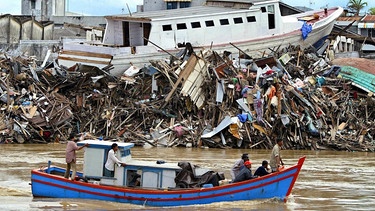 Ein traditionelles Fischerboot fährt am 18.01.2005 im indonesischen Banda Aceh vor einem Trümmerfeld mit einem gestrandeten Schiff vorbei, das von der Tsunami-Welle vom 26.12.2004 zerstört wurde | Bild: picture-alliance/dpa