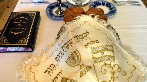 Ein gedeckter Sabbat-Tisch mit dem traditionellen Sabbatbrot (Challot) und einer Karaffe Wein. Das Brot ist mit einem Tuch bedeckt, das mit jüdischen Symbolen und der israelitischen Fahne bestickt ist. | Bild: picture-alliance/dpa