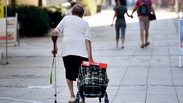 Ältere Dame geht mit Krücke und zieht einen Einkaufstrolley nach. | Bild: picture-alliance/dpa