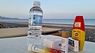 Ein Strand in der Abendstimmung. Im Vordergrund Medikamente, Insektenschutz und Mineralwasser - ein Symbolbild für die Reiseapotheke im Urlaub. | Bild: picture-alliance/dpa
