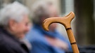 Griff eines Gehstocks, unscharf im Hintergrund ein älterer Mensch. | Bild: picture-alliance/dpa
