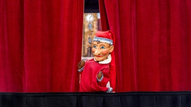 Kasperl schaut hinter einem roten Vorhang hervor. | Bild: picture-alliance/dpa