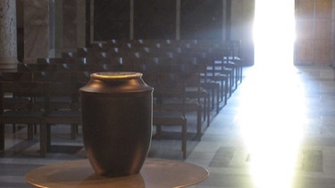 Im Vordergrund eine Urne. Im Hintergrund Kirchenbänke und ein hell erleuchteter Kirchenausgang. | Bild: Gesche Piening