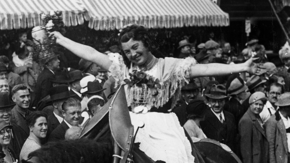 Ein Foto in schwarz-weiß. Eine Frau in Tracht sitzt lachend, mit ausgebreiteten Armen auf einem Wiesn Ross, in der rechten Hand hält sie einen Wiesn-Krug. | Bild: Scherl/Süddeutsche Zeitung Photo