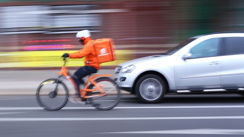 Ein Kurier-Fahrrad-Fahrer fährt knapp vor einem Auto her. Mitgezogene Aufnahme mit längere Belichtungszeit | Bild: picture-alliance/dpa