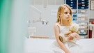Ein kleines Mädchen sitzt auf einem Krankenhausbett. | Bild: stock.adobe.com/rawpixel.com