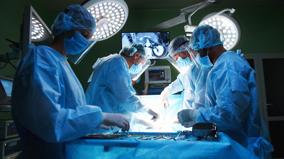 Ein Team aus Ärzten mit sterilen Kitteln, Handschuhen und OP-Hauben führen eine Operation in einem Operationssaal durch. | Bild: stock.adobe.com/ihorvsn