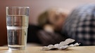Eine junge Frau liegt krank im Bett. Im Vordergrund stehen ein Glas Wasser und ein Blister Tabletten. | Bild: picture alliance/photothek/Ute Grabowsky