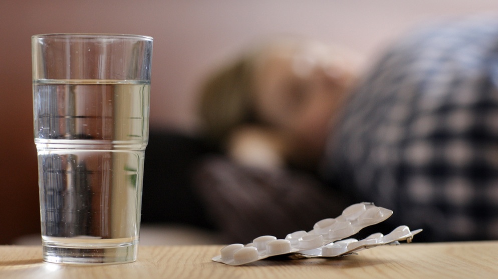 Eine junge Frau liegt krank im Bett. Im Vordergrund stehen ein Glas Wasser und ein Blister Tabletten. | Bild: picture alliance/photothek/Ute Grabowsky