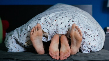 Ein Pärchen liegt im Bett, die Füße schauen aus der Bettdecke hervor.
| Bild: BR/Julia Müller