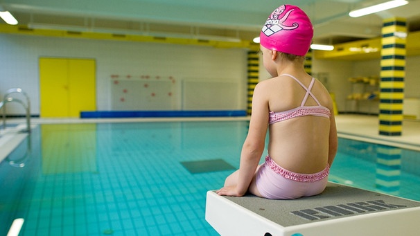 Mädchen sitzt auf Startblock am Beckenrand eines Schwimmbades | Bild: picture-alliance/dpa