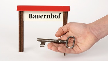 Ein alter rostiger Schlüssel liegt in einer Hand, im Hintergrund ist das Wort Bauernhof zu lesen (Symbolbild) | Bild: picture-alliance/CHROMORANGE/Ernst Weingartner