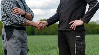 Zwei Männer in landwirtschaftlicher Arbeitskleidung stehen auf einem Getreidefeld und reichen sich die Hand (Symbolbild) | Bild: picture-alliance / Countrypixel | FRP