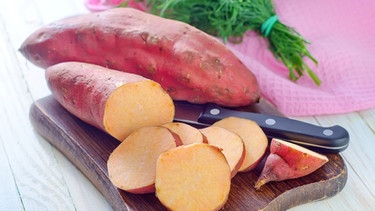 Süßkartoffeln, die auf einem Holzbrett mit einem Messer liegen: die eine Süßkartoffel ist aufgeschnitten, die andere ist im Ganzen | Bild: colourbox.com