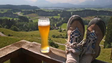 Ein Wanderer genießt mit hochgelegten Füßen und einem Glas Weißbier auf der Terrasse einer Alm bei Seeg (Schwaben) die Aussicht auf das Allgäuer Voralpenland | Bild: picture-alliance/dpa