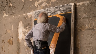 Mann steht auf Stuhl und malt auf Leinwand im Hochformat | Bild: Wolfgang Volz