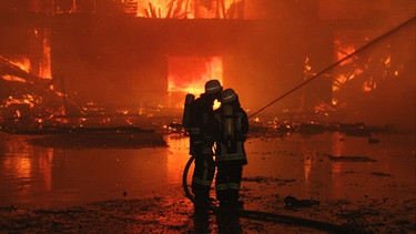 Zwei Feuerwehrmänner stehen vor einem brennenden Haus. | Bild: BR/Georg Barth