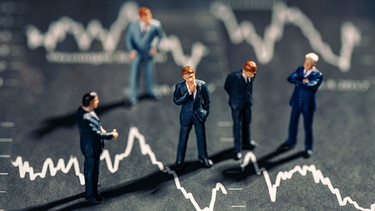 Geschäftsmänner stehen auf Grafiken mit fallenden Börsenkursen. | Bild: picture alliance / Klaus Ohlenschläger | Klaus Ohlenschläger