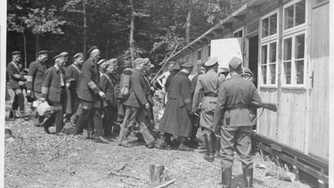Unter Bewachung von SS-Angehörigen werden die ersten Schutzhäftlinge in eine der fertiggestellten Holzbaracken des Lagers gebracht. | Bild: Sammlung Gedenkstätte Buchenwald