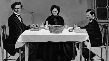 Frau mit schwarzer Haube sitzt am Tisch, links großer Mann, rechts kleiner Mann | Bild: Süddeutsche Zeitung Photo