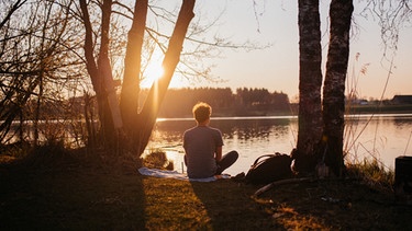 Ein Mann sitzt bei Sonnenuntergang am Ufer eines Sees. | Bild: BR/ Julia Müller