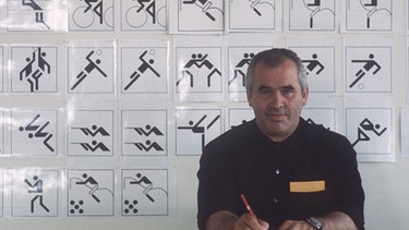 Der Graphiker Otl Aiche, im Hintergrund eine Tafel mit Piktogrammen, die er für die Sportarten der Olympischen Sommerspiele 1972 in München entworfen hat. | Bild: picture-alliance / Sven Simon