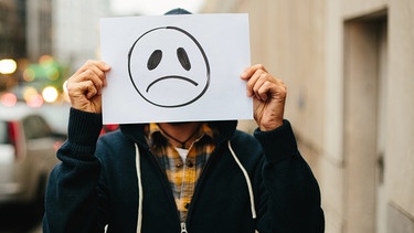 Eine Person hebt ein Schild mit einem traurigem Smiley hoch.  | Bild: BR/Lisa Hinder