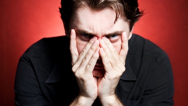 Mann im schwarzen Hemd mit Händen vorm Gesicht blickt angstvoll in die Kamera | Bild: colourbox.com