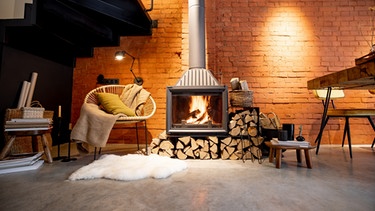 Ein brennender Kamin mit Feuerholz und einem Sessel mit einer Wolldecke. | Bild: stock.adobe.com