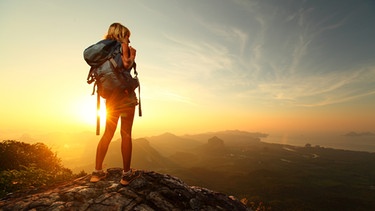Eine Frau steht im Hochsommer auf einem Berg und schaut bei Sonnenaufgang ins Tal hinunter. | Bild: stock.adobe.com/Dudarev Mikhail