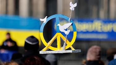 Ein Schild auf einer Demonstration zeigt ein Peace-Zeichen in den Farben der ukrainischen Flagge mit Friedenstauben. | Bild: picture alliance / Geisler-Fotopress | Christoph Hardt/Geisler-Fotopres