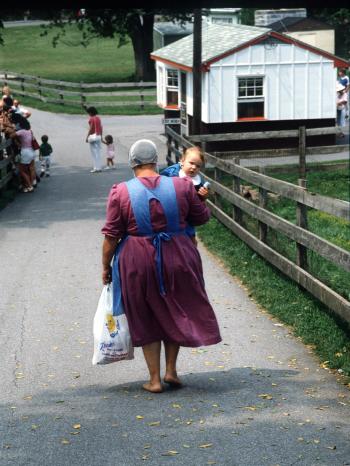 Eine Frau der christlichen Religionsgemeinschaft der Amish geht barfuß mit ihren Einkäufen und einem Säugling auf dem Arm eine Straße entlang. (Aufnahme von 1986).  | Bild: picture-alliance/dpa