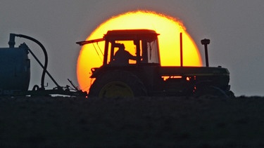 Ein Landwirt fährt auf einem Acker nahe Feldkirchen (Niederbayern) mit seinem Traktor vor der aufgehenden Sonne. | Bild: picture-alliance/dpa