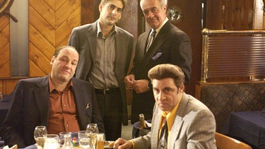 Szene aus den Sopranos: Zwei Männer stehen, zwei Männer sitzen an einem Tisch mit Essen und Wein | Bild: picture-alliance/dpa
