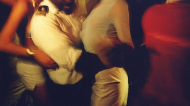 Zwei Frau in engen Kleidern tanzen mit einem uniformierten Mann zwischen sich. Das Bild ist verwackelt und verschwommen. | Bild: picture alliance / PYMCA/Photoshot | Idalina Leandro