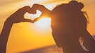 Eine Frau steht vor einem Sonnenuntergang und macht ein Herzzeichen.
| Bild: stock.adobe.com/astrosystem
