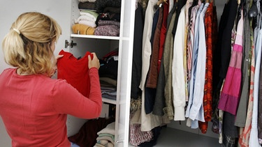 Frau steht vor einem aufgeräumten Schrank und sortiert Kleidung ein | Bild: picture-alliance/dpa