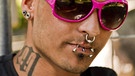Ein junger Mann mit Piercings in Ohr, Nase, Ober- und Unterlippe und einer Tätowierung am rechten Hals. | Bild: picture-alliance/dpa